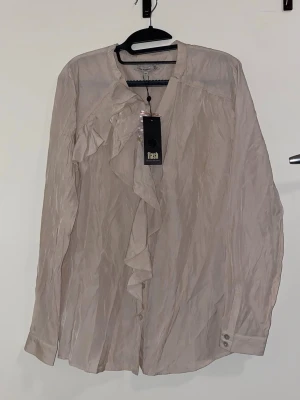 Beige blus med volang  - Stilren beige blus från Flash med långa ärmar och snygg volangdetalj framtill. Blusen har knappar vid ärmsluten och ett lätt glansigt, skrynkligt tyg som ger en cool look. Perfekt för dig som gillar en chic och trendig stil.