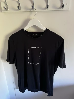 Svart T-shirt från Ted Baker *S* - Snygg T-shirt från Ted Baker med tryckt logotyp i vitt framtill. Klassisk rund halsringning och korta ärmar. Tillverkad i mjuk bomull som känns skön mot huden. Perfekt till jeans eller shorts för en clean look.
