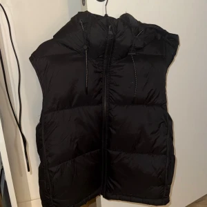 Svart dunväst med huva - Snygg svart dunväst med dragkedja och huva. Västen har en puffig look och justerbara snören vid huvan. Västen är i ett jättefint skick då den endast kommit till användning 1-3 gånger så inga defekter alls den är som ny.