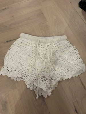 Vita virkade shorts med dragsko - Snygga vita virkade shorts med elastisk midja och dragsko. Shortsens virkade mönster ger en bohemisk känsla och de har en vågig kant nertill. Perfekta för sommardagar eller festivalhäng.
