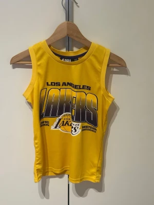 Gul Los Angeles Lakers basketlinne - Snyggt gult basketlinne med Los Angeles Lakers-tryck i lila och vitt framtill. Rund halsringning och ärmlös design. Perfekt för träning eller streetwear.