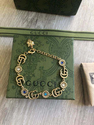Gucci guldarmband med pärlor - Säljer ett lyxigt armband från Gucci i guldfärgad metall med dekorativa pärlor och GG-loggor. Armbandet har blommiga detaljer med vita och blå pärlor samt justerbar kedja och ett lejonhuvud vid låset. Perfekt accessoar för dig som gillar statement-smycken.