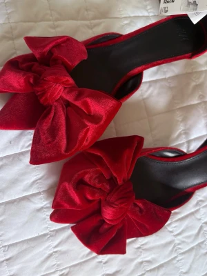 Röda pumps med stor rosett H&M - Säljer ett par röda pumps från H&M i sammetsliknande material. Skorna har öppen häl och en stor, iögonfallande rosett framtill. Klacken är låg och modellen är elegant med en trendig twist. Perfekta för dig som vill sticka ut med färg och detaljer.