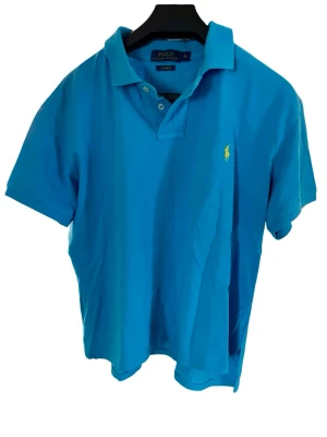 Blå pikétröja från Polo Ralph Lauren (XL) - Snygg blå pikétröja från Polo Ralph Lauren i storlek XL med klassisk krage, korta ärmar och gul broderad logga på bröstet. Perfekt för en avslappnad och stilren look. Tillverkad i mjuk bomull för extra komfort. I bra skick utan fläckar eller nämnvärda tecken på slitage. 