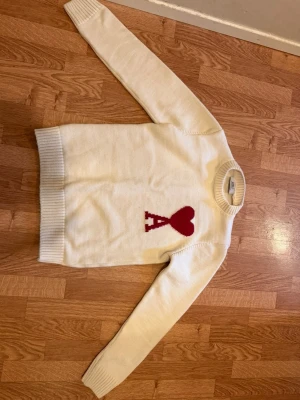 Vit stickad tröja från Ami - Vit stickad tröja från Ami Alexandre Mattiussi med röd hjärt- och A-detalj på bröstet. Tröjan har rund halsringning och ribbstickade muddar vid ärmslut och nederkant. Perfekt för dig som gillar stilrena och trendiga plagg. Pris kan diskuteras 