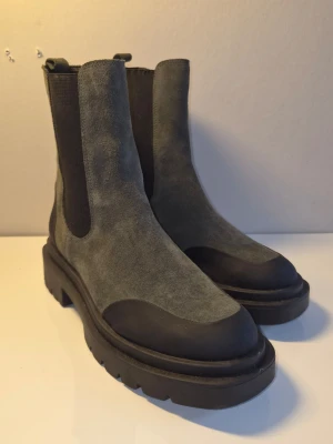 Grå chunky Chelsea boots i mocka - Säljer ett par grå Chelsea boots med chunky sula och svarta detaljer. Skorna är i mocka med elastiska partier på sidorna och grov yttersula i gummi. Perfekta för dig som vill ha en cool och trendig look till hösten eller vintern.