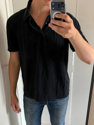 Svart piké från Baldessarini - Säljer en svart kortärmad skjorta från Baldessarini Hugo Boss. Skjortan har klassisk krage och knappar framtill. Tillverkad i ett mjukt material som känns skönt mot huden. Perfekt till jeans eller chinos för en clean look.