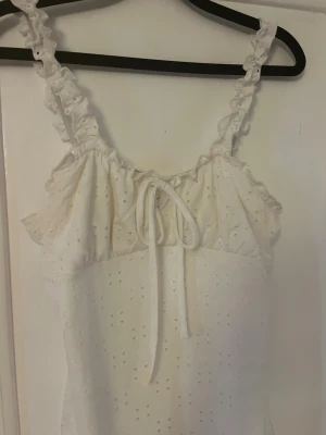 Vit kort klänning med volang och knyt - Söt vit kort klänning med broderade detaljer, volang vid axelbanden och knyt framtill. Klänningen har ärmlös design och romantisk känsla, perfekt för varma dagar. Tillverkad i bomull för en luftig och bekväm känsla. Är i L men skulle även passa en S pga hur stretchig den är.