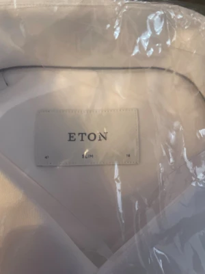 Vit slim fit skjorta från Eton - Klassisk vit skjorta från Eton i slim fit-modell. Skjortan har knappar framtill och vid manschetterna, samt en stilren krage. Tillverkad i ett mjukt och exklusivt bomullstyg som känns lyxigt mot huden. Perfekt för dig som gillar en clean och tidlös look.