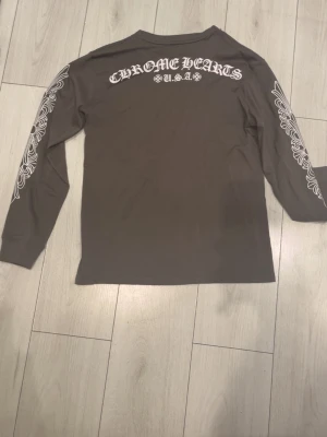 Långärmad topp från Chrome Hearts - Mörkgrå långärmad topp från Chrome Hearts med vita grafiska tryck längs båda ärmarna och logga på bröstet. Stort Chrome Hearts-tryck på ryggen. Skön passform och cool design som sticker ut.