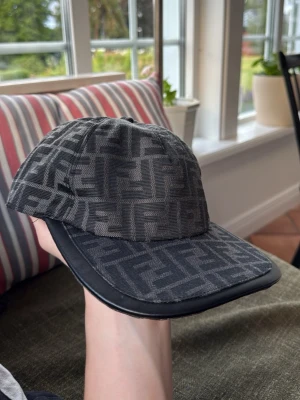 Svart keps med Fendi-logga - Säljer en svart keps från Fendi med det klassiska FF-monogrammet i grått över hela kepsen. Kepsen har böjd skärm med svart kant i läderimitation och justerbar passform. Perfekt för dig som vill ha en stilren och lyxig accessoar.