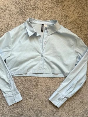 Ljusblå croppad skjorta från H&M - Snygg ljusblå croppad skjorta från H&M Divided med klassisk krage och långa ärmar. Skjortan har knappar framtill och är tillverkad i mjuk bomull. Perfekt för en trendig och avslappnad stil.