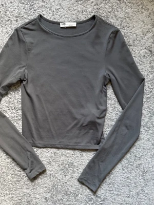 Mörkgrå långärmad crop top - Snygg mörkgrå långärmad crop top från STR Teen. Tröjan har en enkel och stilren design med rund halsringning och är tillverkad i ett mjukt, stretchigt material som sitter skönt på kroppen. Perfekt att matcha med höga jeans eller kjol.