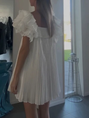 Vit plisserad kortklänning med volang - Supersöt vit kortklänning med plisserad kjol och stora volangärmar. Klänningen har en rak passform och fyrkantig ringning, vilket ger en trendig och luftig look. Perfekt för sommarens alla tillfällen!