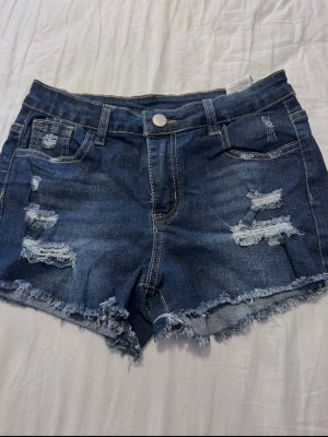 Blå denimshorts med slitningar från SHEIN - Säljer ett par blå denimshorts från SHEIN med råa kanter och slitna detaljer framtill. Shortsen har fem fickor, bälteshällor och knappstängning. Perfekta för sommaren och ger en avslappnad streetstyle-look.