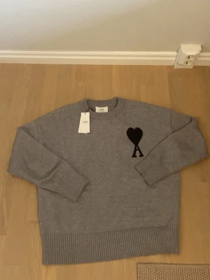 Ami Paris grå tröja  - Snygg grå stickad tröja med svart hjärta och bokstaven A på bröstet. Tröjan har ribbade muddar vid ärmslut och nederkant samt en klassisk rund halsringning. Perfekt för dig som gillar stilrena och trendiga plagg.