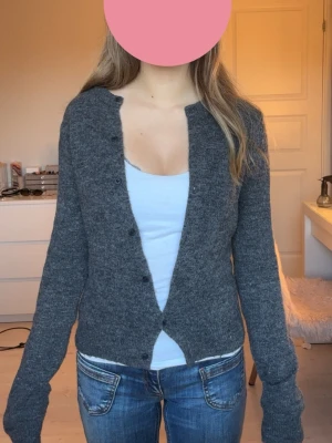 Mörkgrå stickad kofta - Mysig mörkgrå stickad kofta med rund halsringning och knäppning framtill. Koftan har långa ärmar och en enkel, klassisk design som passar till det mesta. Perfekt att slänga över en topp eller t-shirt för extra värme.
