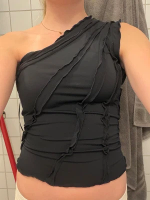 Svart oneshoulder topp med råa sömmar - Säljer en svart oneshoulder topp med råa, dekorativa sömmar och asymmetrisk skärning. Toppen är tight och croppad, perfekt för dig som vill ha en edgy och trendig look. Materialet känns mjukt och stretchigt.