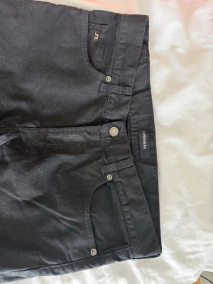 Svarta jeans från J.Lindeberg - Snygga svarta jeans från J.Lindeberg med klassisk femficksmodell och raka ben. Jeansen har silverfärgade detaljer och diskret JL-logga vid fickan. Materialet är slitstarkt och passar perfekt till en trendig vardagslook.