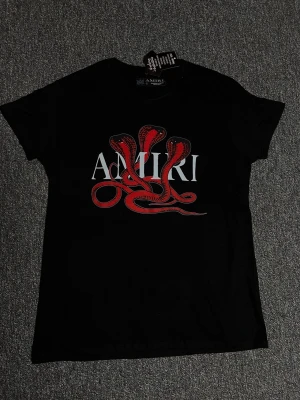 Svart Amiri t-shirt med röd orm - Snygg svart t-shirt från Amiri med ett stort tryck av en röd orm och vit Amiri-logga på bröstet. T-shirten har korta ärmar och rund hals. Perfekt för dig som vill sticka ut med en cool och edgy look.