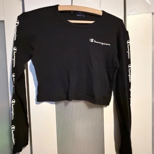 Svart croppad långärmad topp Champion - Svart croppad långärmad topp från Champion med logga tryckt på bröstet och längs båda ärmarna. Rund halsringning och normal passform. Perfekt för en sportig och avslappnad stil.