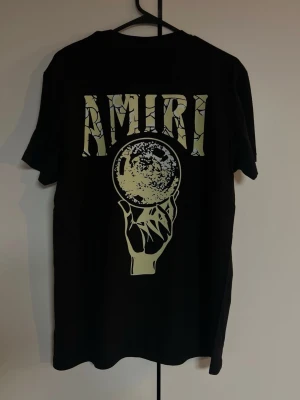 Svart Amiri t-shirt med tryck - Säljer en svart t-shirt från Amiri med stort grafiskt tryck på ryggen. Trycket visar en hand som håller en klotformad kristallkula och texten 'AMIRI' i ett sprucket mönster ovanför. Perfekt för dig som gillar streetwear och unika prints.