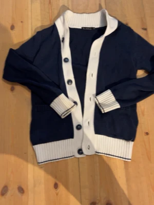 Marinblå cardigan från bondolid - Stilren marinblå cardigan med breda vita ribbade muddar och knappslå. Tröjan har långa ärmar och stora knappar framtill. Perfekt för en avslappnad men ändå snygg look.