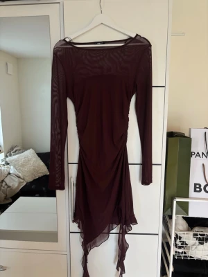Vinröd mesh klänning - Vinröd klänning i mesh med asymmetrisk och volangig nederdel. Klänningen har lång ärm och transparent tyg som ger en edgy look. Perfekt för dig som vill sticka ut med en unik siluett och coola detaljer. Köpt på New Yorker i storlek L, passar bra på en M också