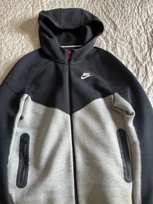Nike hoodie svart och grå XS - Nike hoodie i storlek XL 158-170 barnstorlek, med svart ovandel och grå nederdel. Dragkedja framtill, två fickor med svarta dragkedjor och klassisk Nike-logga på bröstet. Tillverkad i mjukt och stretchigt material, perfekt för chill eller träning. Snygg kontrastdesign och huva.