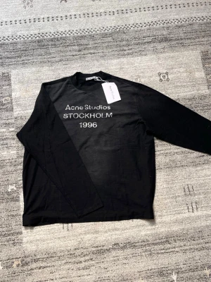 Svart Acne Studuios tröja - Svart långärmad topp från Acne Studios med tryck 'Acne Studios STOCKHOLM 1996' i vitt framtill. Klassisk rund halsringning och mjukt material som känns skönt mot huden. Perfekt för dig som gillar minimalistisk och stilren design.