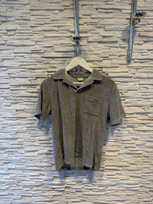 Grå t-shirt med krage och bröstficka - Säljer en grå t-shirt med klassisk krage och en bröstficka. T-shirten har korta ärmar och är tillverkad i ett mjukt material som känns skönt mot huden. Perfekt för en avslappnad och stilren look.