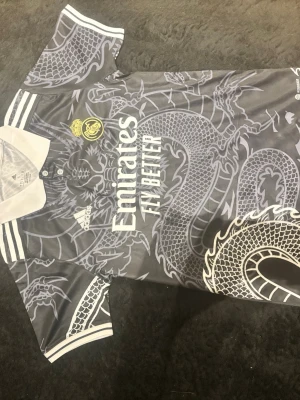 Real Madrid x Adidas draktröja S - Säljer en grå och vit Real Madrid matchtröja från Adidas med stort drakmönster, klubbmärke och sponsortryck på bröstet. Tröjan har vit krage, korta ärmar och är tillverkad i återvunnet material. Perfekt för dig som älskar fotboll och unik design.