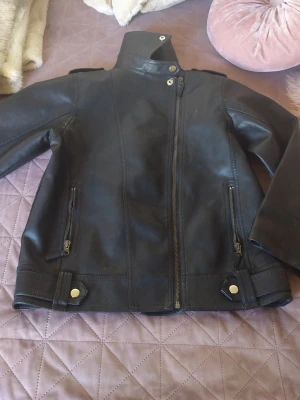 Svart skinnjacka med ståkrage - Snygg svart skinnjacka med asymmetrisk dragkedja, ståkrage och detaljer i guld. Två framfickor med dragkedja och dekorativa axelremmar. Perfekt för dig som vill ha en klassisk men edgy look.