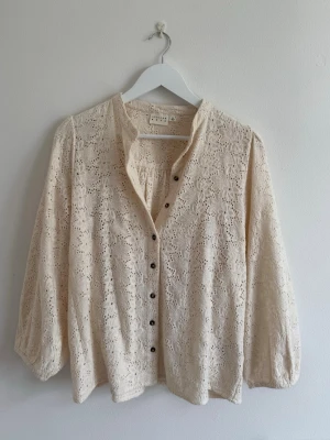 Krämvit spetsblus med knappar - Söt krämvit blus från Atelier Reve i broderad spets med v-ringning och dekorativa svarta knappar framtill. Blusen har lång ärm med lätt puff och resår vid ärmslut. 