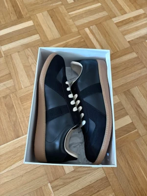 Maison Margiela gats storlek 45 - Svarta sneakers från Maison Margiela med beige detaljer och vita snören. Skorna har en klassisk låg siluett, ovandel i mocka och läder samt en gummisula i ljusbrunt. Snygg retrostil som passar till många outfits.