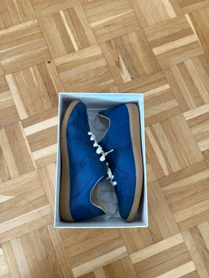 Blå  Maison Margiela gats storlek 43 - Snygga blå sneakers från Maison Margiela med vita skosnören och beige gummisula. Skorna har en mix av mocka och läderdetaljer, klassisk låg silhuett och diskret logga på plösen. Perfekta för dig som gillar stilrena och trendiga sneakers.