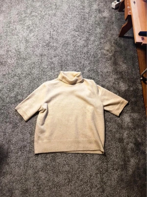 Beige stickad polotröja kort ärm - Liten i storleken!!  Stickad beige polotröja med korta ärmar och avslappnad passform. Tröjan har ribbade kanter och hög krage som ger en stilren look. Perfekt att matcha med jeans eller kjol för en enkel men snygg outfit.