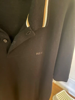 Svart pikétröja från BOSS - Klassisk svart pikétröja från BOSS med korta ärmar och krage med vita och beige detaljer. Tröjan har knappar framtill och diskret BOSS-logga på bröstet. Perfekt för en clean och stilren look.