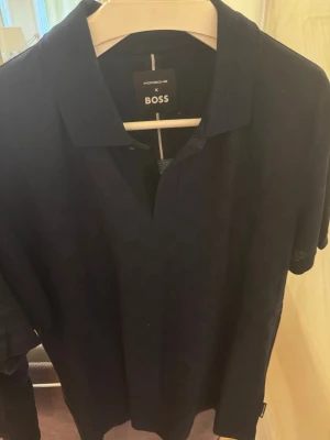 Mörkblå pikétröja Porsche x BOSS - Snygg mörkblå pikétröja från Porsche x BOSS med klassisk krage och kort ärm. Perfekt för en stilren och sportig look. Tröjan har diskret logga i nacken och är tillverkad i mjukt material.