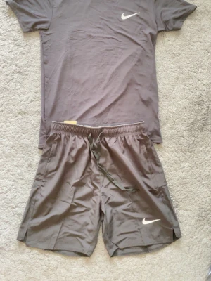 Nike träningsset t-shirt och shorts - Säljer ett sportigt träningsset från Nike med en grå t-shirt och matchande grå shorts, båda med den klassiska vita Nike-loggan. Materialet är lätt och snabbtorkande, perfekt för träning eller löpning. T-shirten har rund hals och korta ärmar, shortsen har dragsko i midjan.