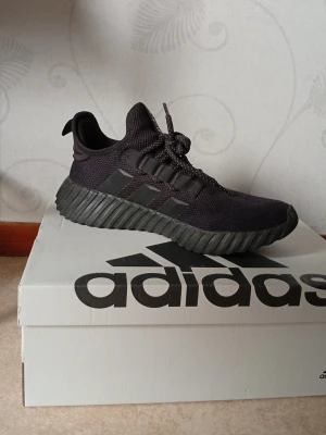 Svarta Adidas skor - Snygga svarta sneakers från Adidas med klassiska tre ränder på sidan. Ovandelen är i meshmaterial för bra andningsförmåga och sulan är kraftig och räfflad för extra grepp. Skorna har snörning och en sportig, modern look som passar till det mesta.
