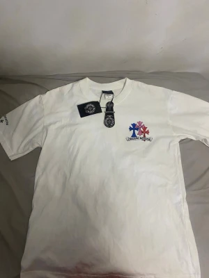 Vit t-shirt från Chrome Hearts - Vit t-shirt från Chrome Hearts med tryck av blått och rött kors på bröstet samt logga under. Klassisk rund hals och korta ärmar. Tillverkad i mjuk bomull och har märkets taggar kvar. Perfekt för dig som gillar streetwear och vill sticka ut.