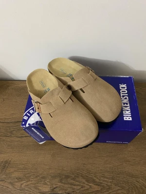 Beige Birkenstock Boston sandaler - Säljer ett par beige Birkenstock Boston sandaler i mocka med klassisk slip-in design och justerbart spänne i sidan. Yttersulan är svart och har ett vågigt mönster för bra grepp. Perfekta för en avslappnad och trendig stil.