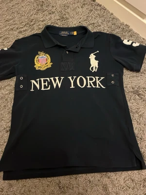 Ralph Lauren piké - Säljer en mörkblå pikétröja från Polo Ralph Lauren med broderad logga, emblem och texten 'NEW YORK' på bröstet. Tröjan har korta ärmar, krage och knappar vid halsen. Slim fit-modell som passar perfekt till en sportig och stilren look. Står att det är storlek S men skulle säga att den passar bättre som XS. Hör av er vid frågor eller intresse!😇