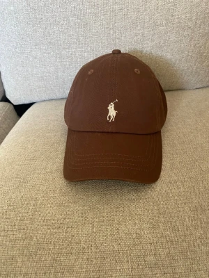 Brun keps från Polo Ralph Lauren - Snygg brun keps från Polo Ralph Lauren med klassisk broderad logga framtill och texten 'POLO' baktill. Justerbart spänne i metall bak för perfekt passform. Tillverkad i bomull och har en böjd skärm.
