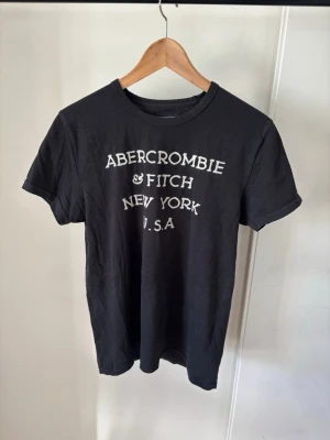 Svart Abercrombie & Fitch t-shirt - Svart t-shirt från Abercrombie & Fitch med vit text på bröstet där det står 'Abercrombie & Fitch New York U.S.A'. Klassisk rund halsringning och korta ärmar. Perfekt för en avslappnad och stilren look.