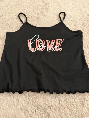 Svart linnetopp/pyjamastopp - Snygg svart linnetopp (pyjamasbyxor) med tunna axelband och vågig kant nertill. Framtill står det 'LOVE' i rosa och vitt tryck. Perfekt för en trendig och avslappnad stil! - Helt oanvänd 