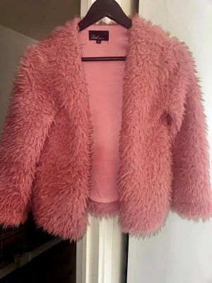 Rosa fuskpälsjacka XS Zoul Edition - Supersnygg rosa teddyjacka med lång luddig pälsimitation och öppen front. Jackan är i använt men fint skick. Materialet är mjukt och fluffigt, vilket gör den extra skön att bära.