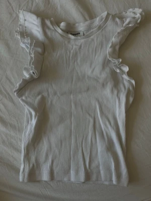 Vit ribbad linnetopp med volang - Supersöt vit ribbad linnetopp från Kappahl med volangdetaljer vid ärmhålen. Toppen är ärmlös och har en tight passform som framhäver figuren. Perfekt att styla med jeans eller kjol för en fräsch look.