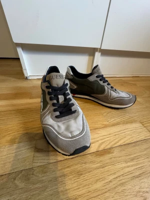 Guess sneakers i grått och olivgrönt - Snygga sneakers från Guess i grått och olivgrönt med svarta detaljer. Skorna har svarta snören, vit sula och logga på plösen. Materialmix av mocka och textil ger en modern look. Perfekta för dig som vill ha stilrena och bekväma sneakers.
